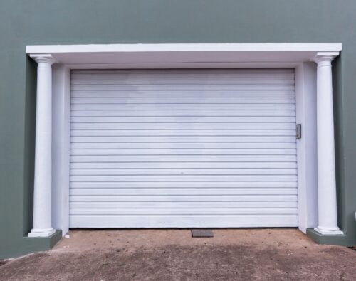 A closed white garage door with vertical ridges, framed by two white columns against a green wall. Concrete driveway in front.
