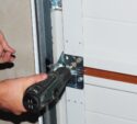 A person uses a power drill to secure a metal bracket onto a white garage door.