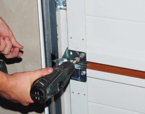 A person uses a power drill to secure a metal bracket on a white garage door.