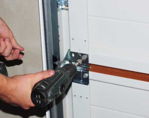 A person uses a power drill to secure a metal bracket onto a white garage door.