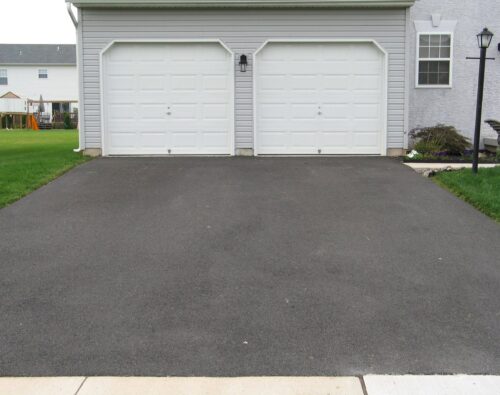 A double garage with two white doors and a freshly paved asphalt driveway in front of a light gray house.