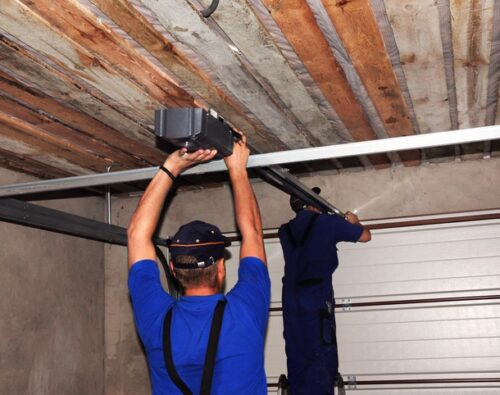 Two workers in blue uniforms install an automatic garage door opener on the ceiling of a garage with an unfinished wooden roof.