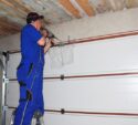 Contractor standing on a ladder using a power drill to install or repair a garage door inside a garage with exposed wooden ceiling.