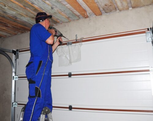 Contractor standing on a ladder using a power drill to install or repair a garage door inside a garage with exposed wooden ceiling.