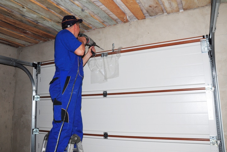 Contractor standing on a ladder using a power drill to install or repair a garage door inside a garage with exposed wooden ceiling.