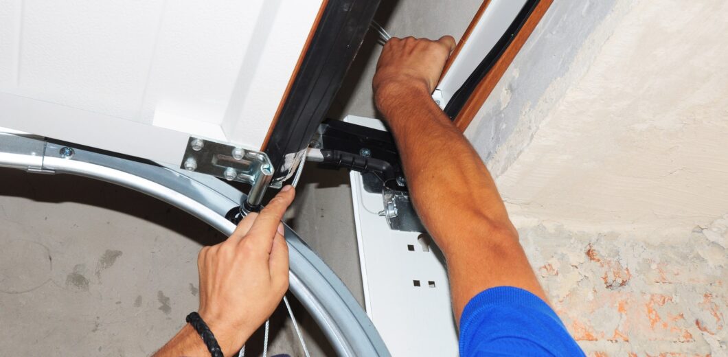 A person wearing a blue shirt is installing or repairing the mechanism of a white garage door using tools.