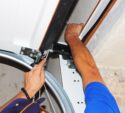 A person wearing a blue shirt is installing or repairing the mechanism of a white garage door using tools.