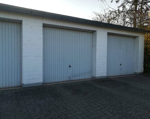 Three closed, white, single-car garage doors with vertical panels are set in a white brick building on a paved driveway.