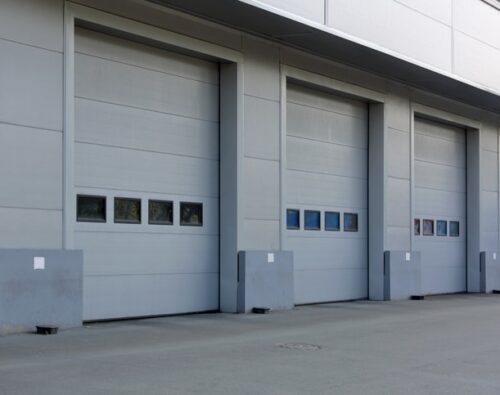 Four closed industrial garage doors on a gray building, each with a row of rectangular windows and protective barriers at the base.