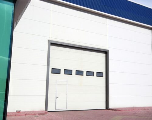 Large white industrial garage door with five horizontal windows and a small pedestrian door at the bottom, set in a warehouse exterior with blue trim and red brick pavement.