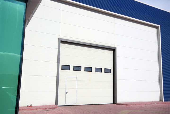 Large white industrial garage door with five horizontal windows and a small pedestrian door at the bottom, set in a warehouse exterior with blue trim and red brick pavement.