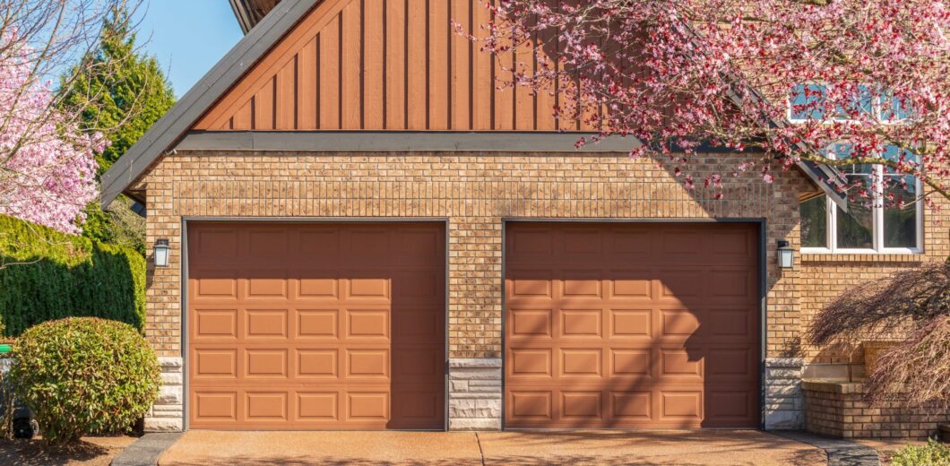 Two closed brown garage doors on a brick house, with blooming pink trees framing the driveway on a sunny day.