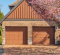 Two closed brown garage doors on a brick house, with blooming pink trees framing the driveway on a sunny day.