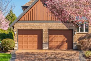Two closed brown garage doors on a brick house, with blooming pink trees framing the driveway on a sunny day.