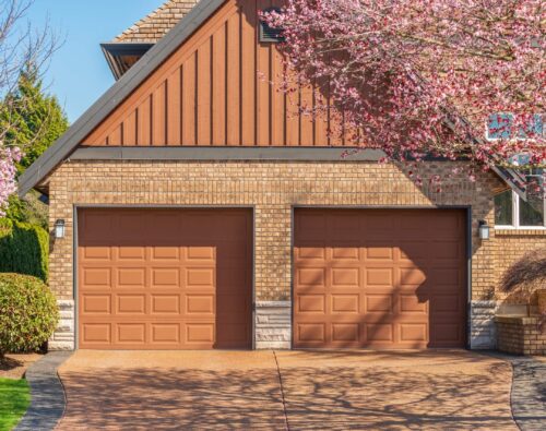 Two closed brown garage doors on a brick house, with blooming pink trees framing the driveway on a sunny day.