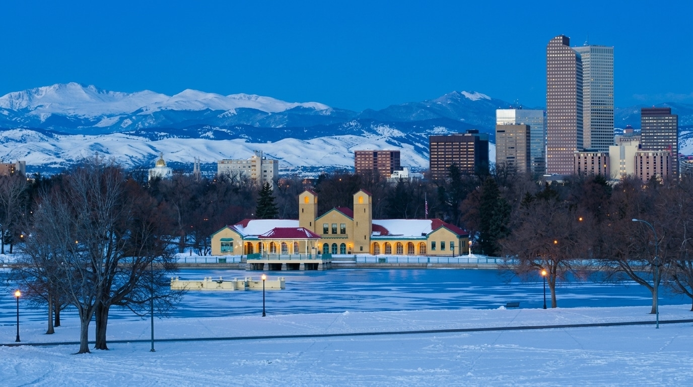 City skyline with snow-covered park and mountains in the background at dusk; a large yellow building sits by a frozen lake in the foreground, capturing the serene winter charm of Aurora, CO.