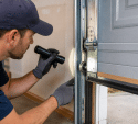 Garage door technician wearing gloves uses a flashlight to inspect the side track, rollers, and hinges of a partially open residential garage door.